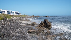 Een hotel aan de kust met een rotsachtige oever, een helderblauwe lucht en golven die op de rotsen breken.
