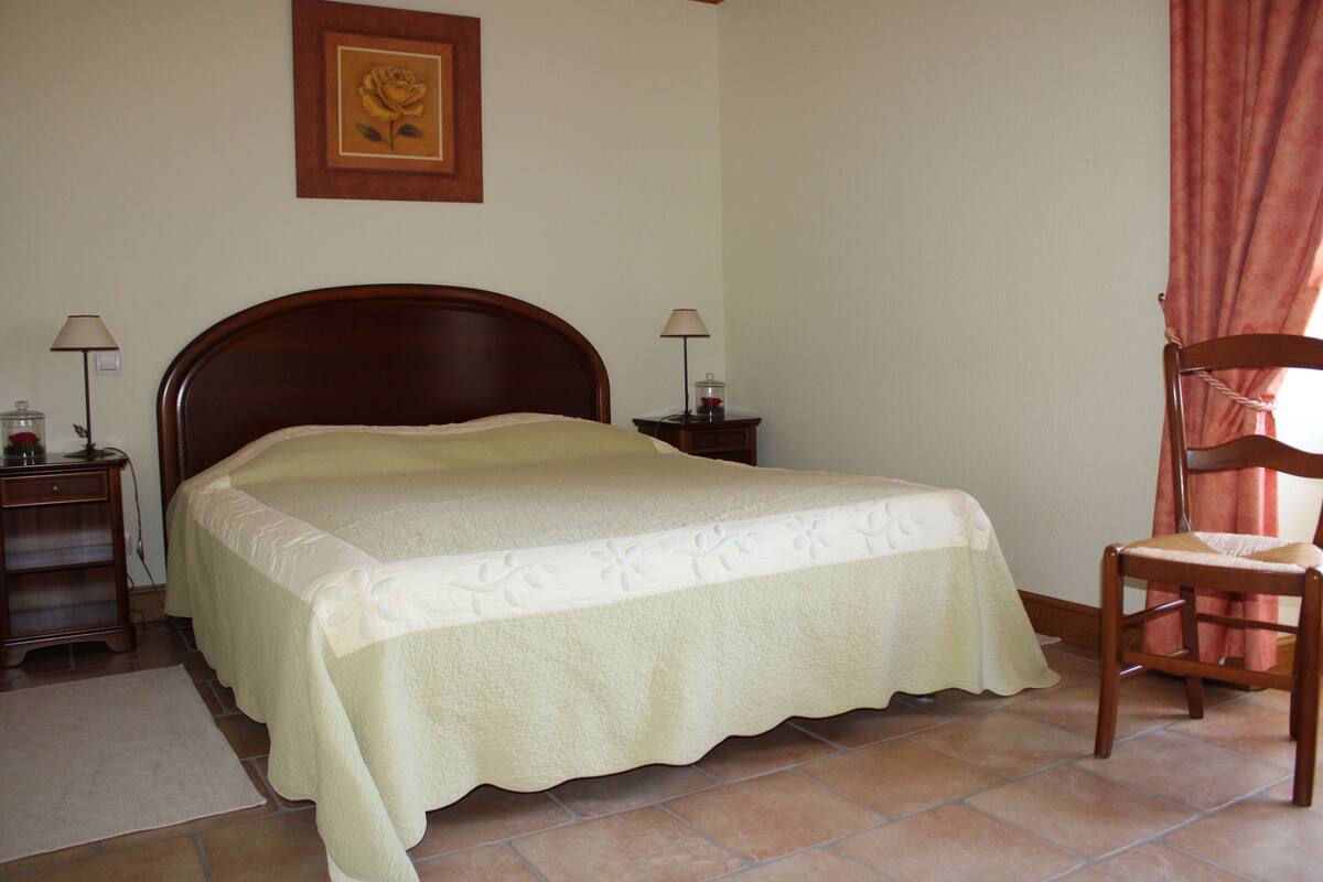 Una cama bien hecha con una colcha floral, dos mesitas de noche con lámparas, una silla de madera y un cuadro en la pared.