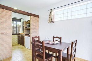 A dining area with a wooden table and chairs, a kitchen in the background, and a window with curtains.