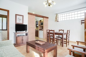 A living room with a TV, wooden furniture, a dining area, and a kitchen in the background.