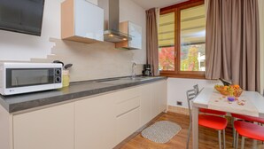 A kitchen with a microwave, cabinets, a window with curtains, a table with a fruit bowl, and red chairs. - Holiday Apartment "GeoResort B6" with Panoramic Views and Shared Pool (Tremosine)