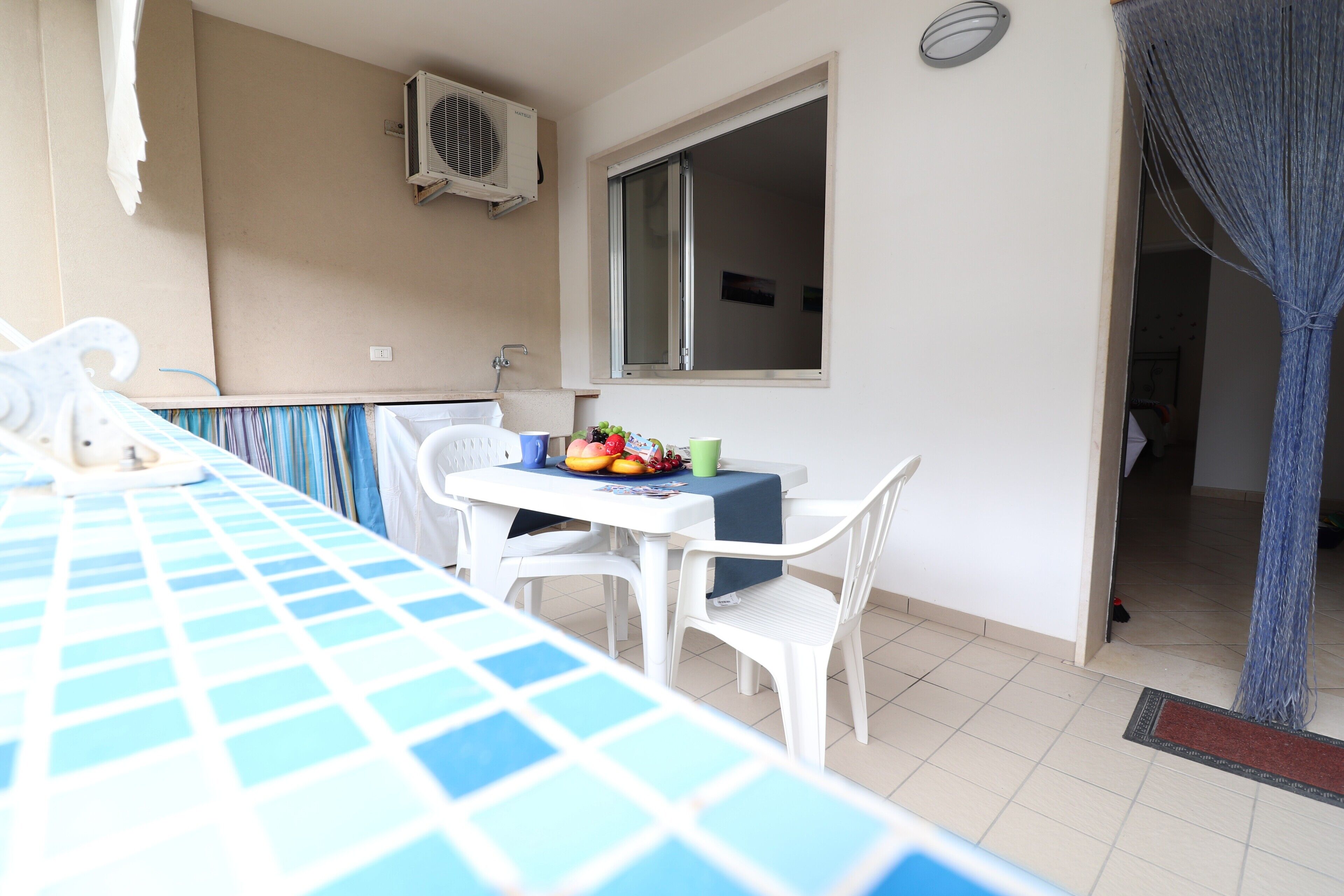 Ein Balkon mit Tisch für zwei Personen, einem Stuhl, einem kleinen Tisch mit Früchten und einer grünen Tasse, einem Fenster und einer Klimaanlage.