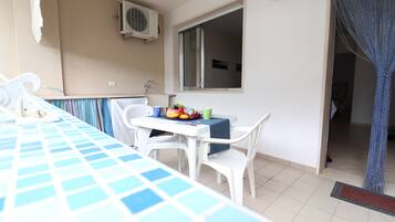 Ein Balkon mit Tisch für zwei Personen, einem Stuhl, einem kleinen Tisch mit Früchten und einer grünen Tasse, einem Fenster und einer Klimaanlage.