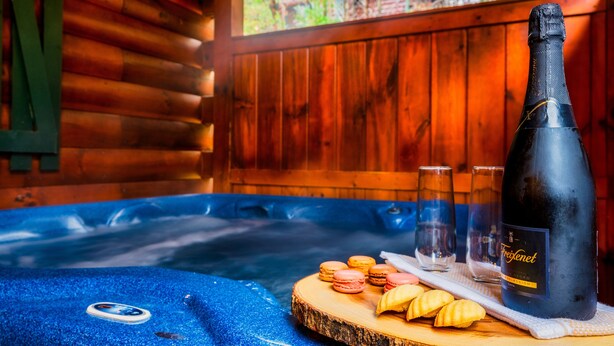 Una cabaña de madera con un jacuzzi, una botella de vino, copas y macarons sobre una tabla de madera.
