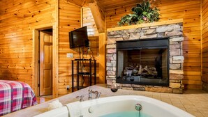 Interior de una cabaña de madera con chimenea de piedra, televisión y bañera.