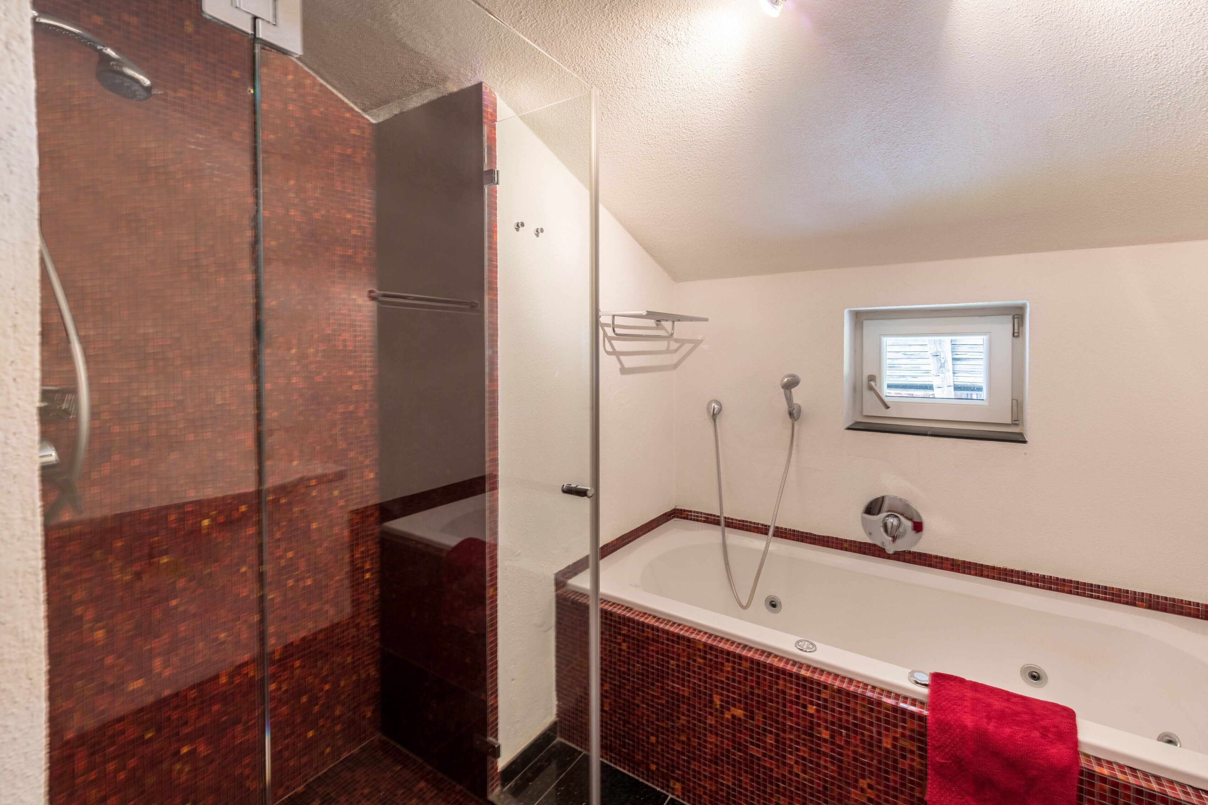 Ein Badezimmer mit Duschkabine aus Glas, einer Badewanne und einem roten Handtuch sowie einem Fenster über der Wanne.
