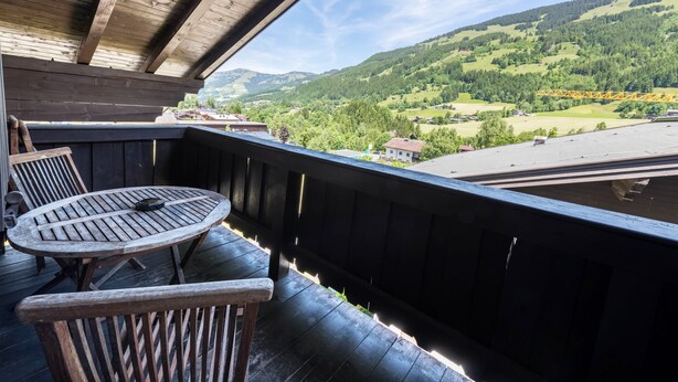 Een houten terras met een tafel en stoelen, met uitzicht op een vallei met huizen en een brug.