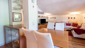 Een moderne woonkamer met een houten eettafel, witte stoelen, een glazen kast met servies, een televisie en een bank met rode kussens.