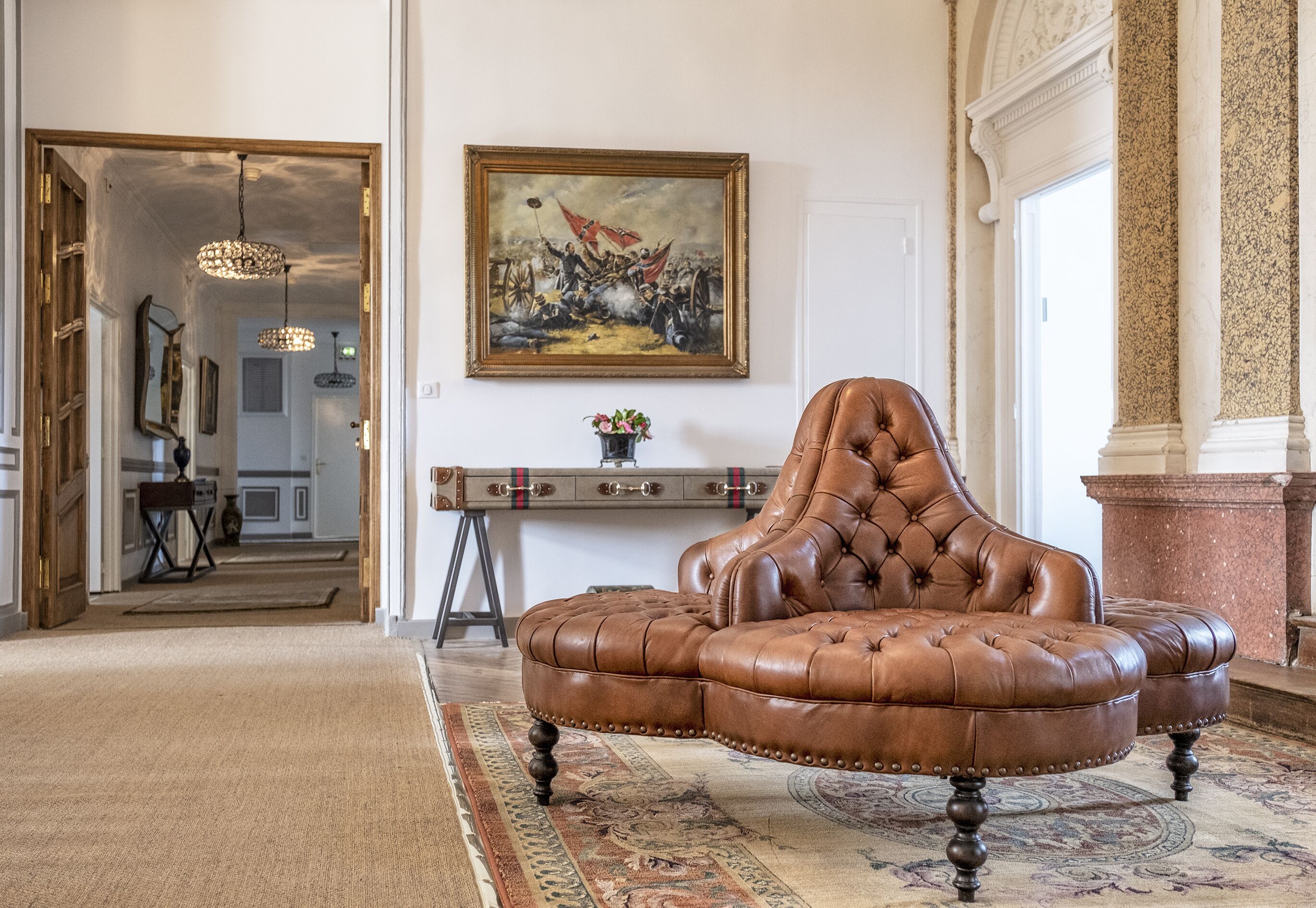 A brown leather armchair in a spacious room with a large painting on the wall, a wooden desk, and a patterned rug.
