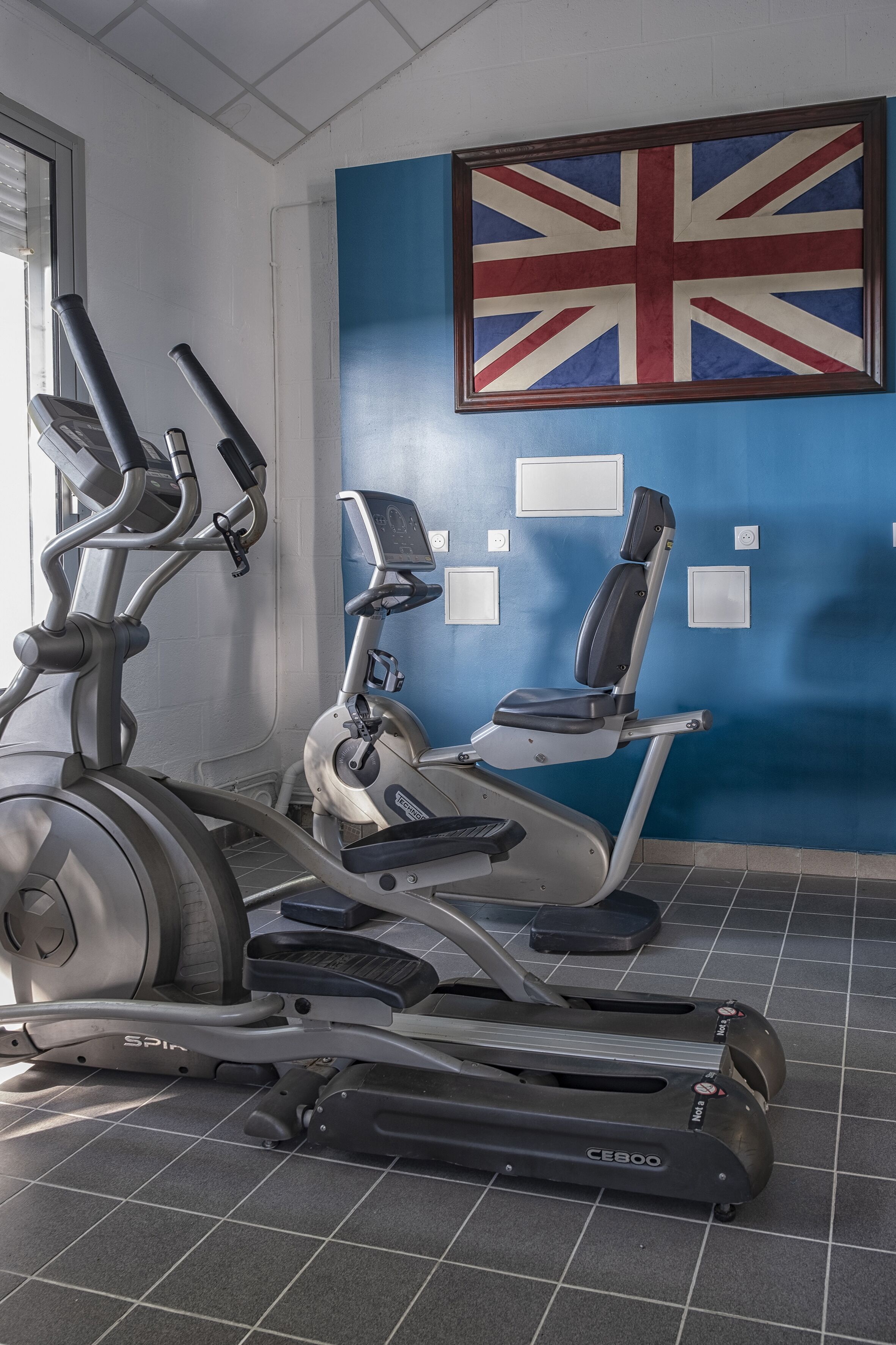 Ein Fitnessraum mit Ergometer und Laufband, eine blaue Wand mit einer Union-Jack-Flagge und ein Fenster mit Jalousien.