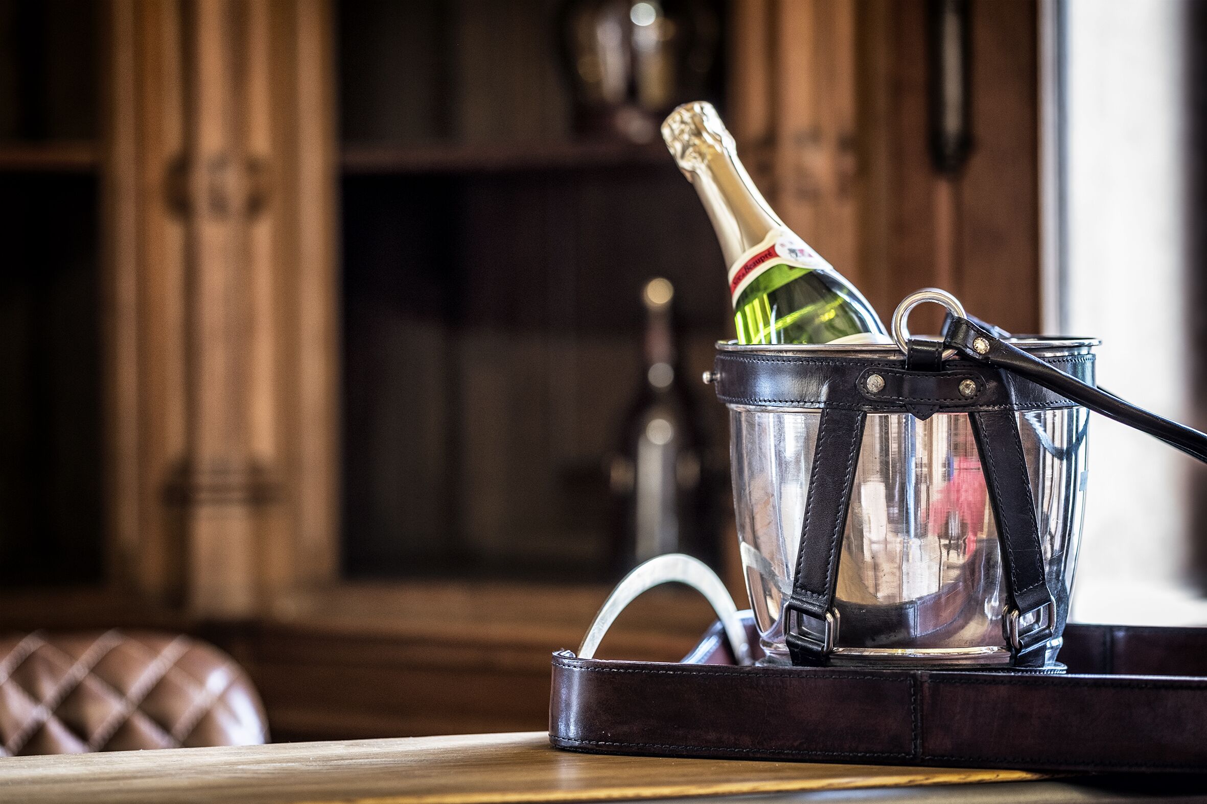 A bottle of champagne in a clear glass container with a black strap, placed on a wooden table in a room with wooden furniture and a dark background.