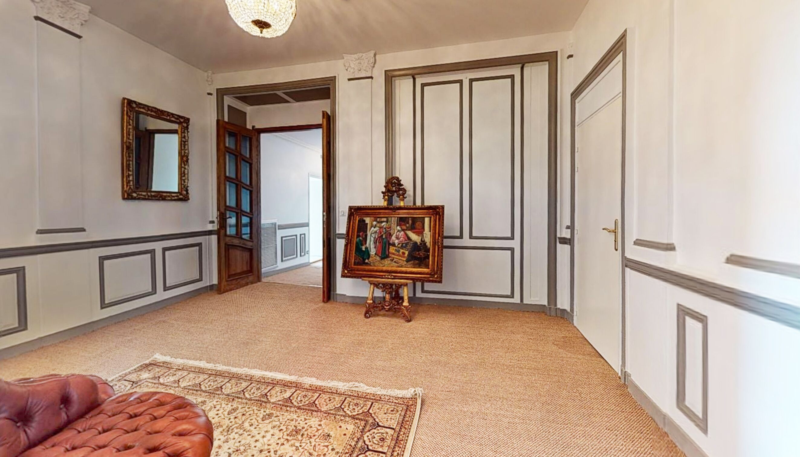 A hallway with a patterned carpet, a framed painting on an easel, and a brown leather ottoman.