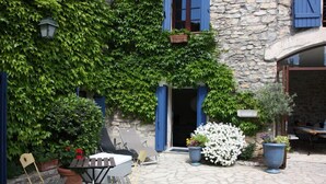 A stone building with blue shutters, a small outdoor seating area with a table and chairs, and potted plants. - Chambre d'hôtes Bleus Matines (Caveirac)
