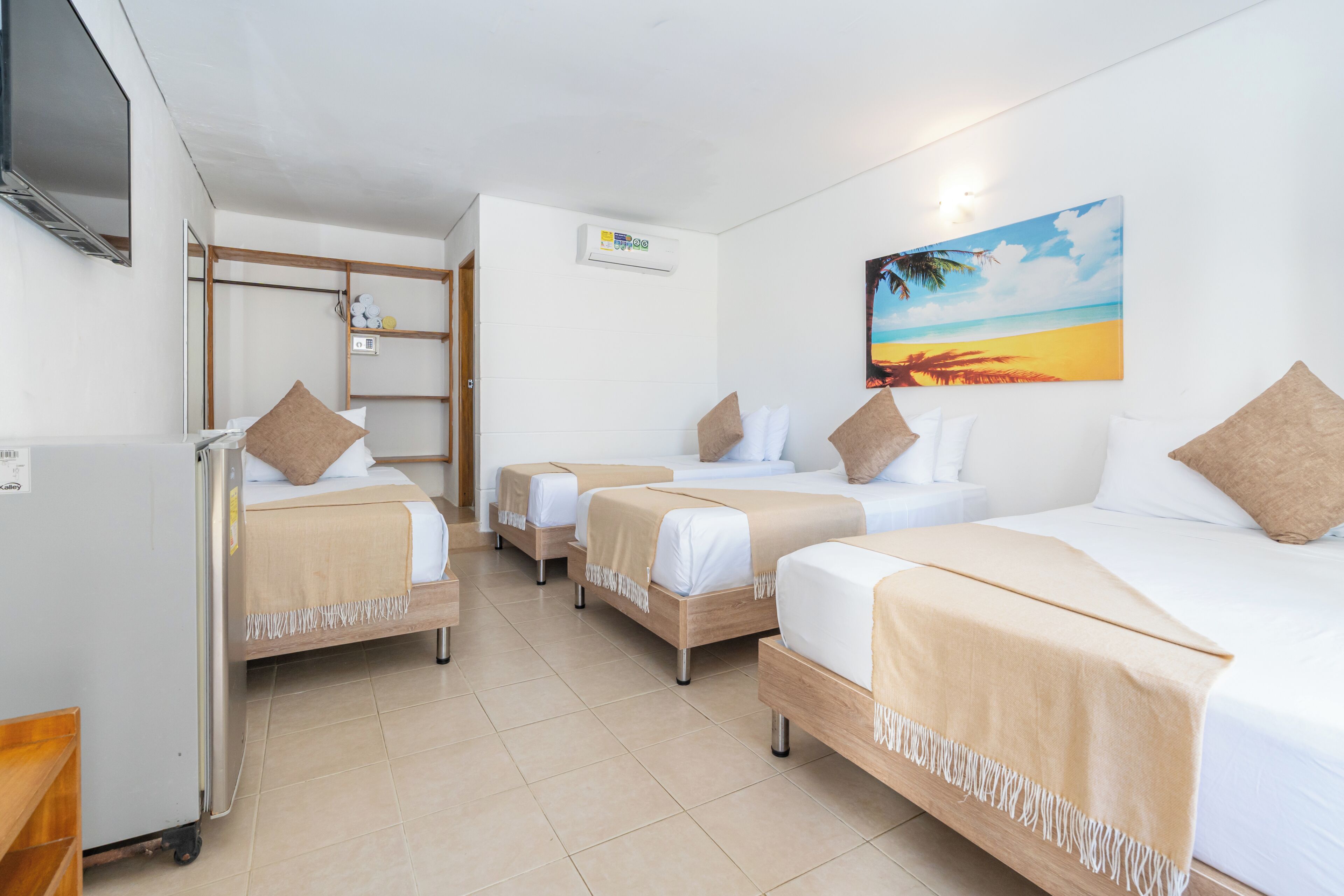 A hotel room with three beds, a TV, a refrigerator, and a painting of a beach scene.