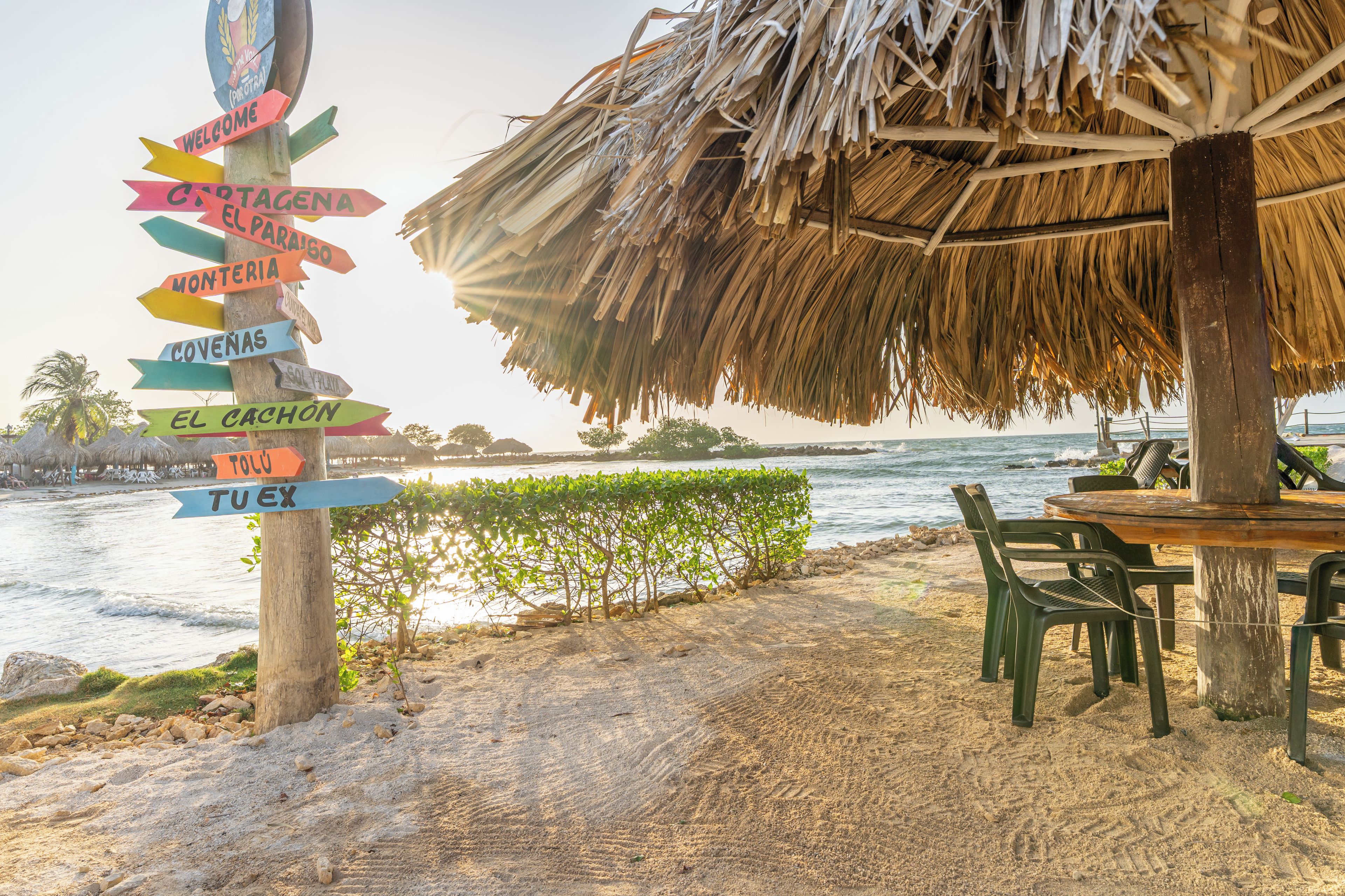 Escena en la playa con un poste de madera con flechas direccionales, una estructura con techo de paja y una mesa con sillas bajo una palmera.