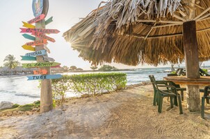 Escena en la playa con un poste de madera con flechas direccionales, una estructura con techo de paja y una mesa con sillas bajo una palmera.