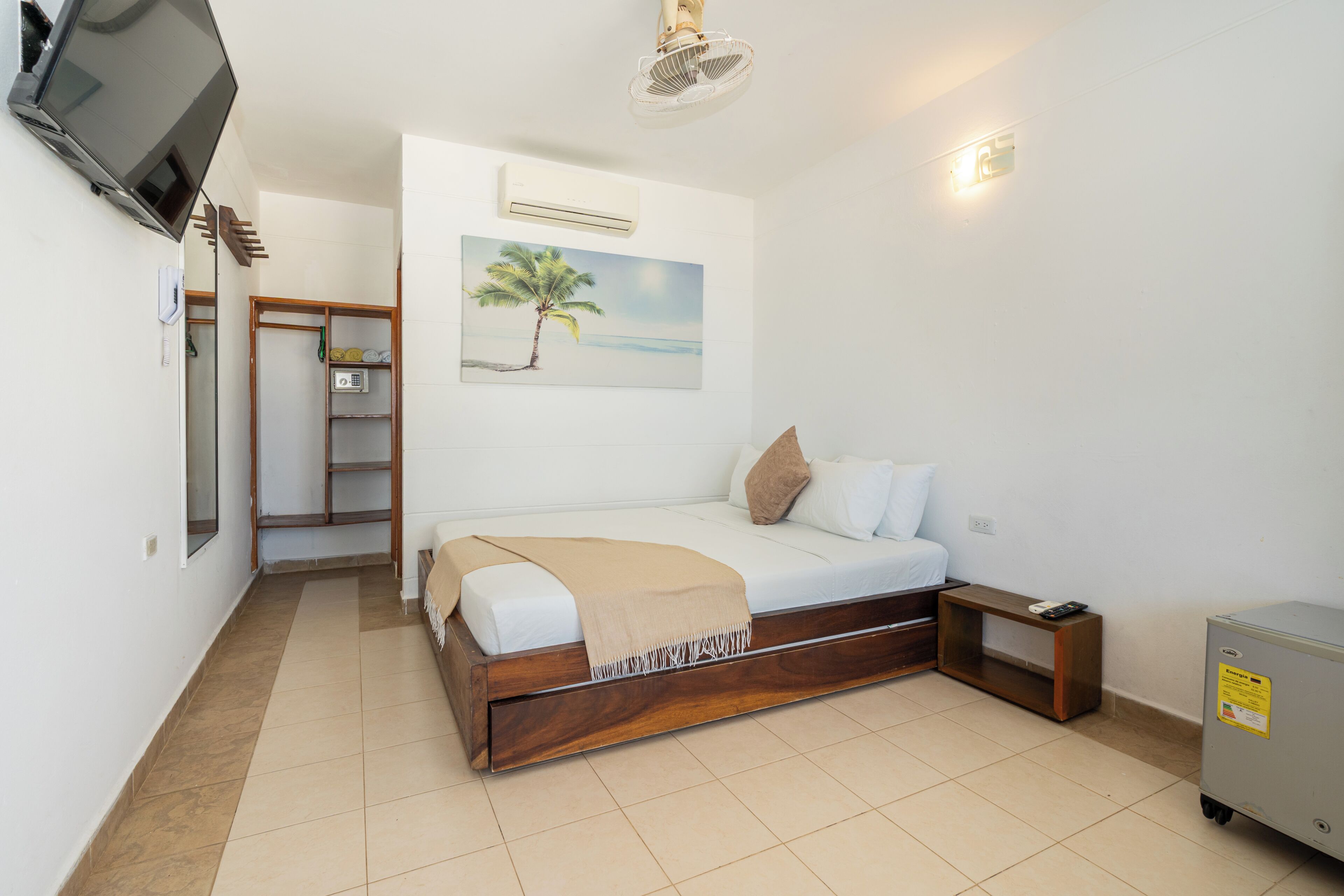 Un dormitorio con cama, televisor, un cuadro de una palmera, armario y una pequeña mesa.