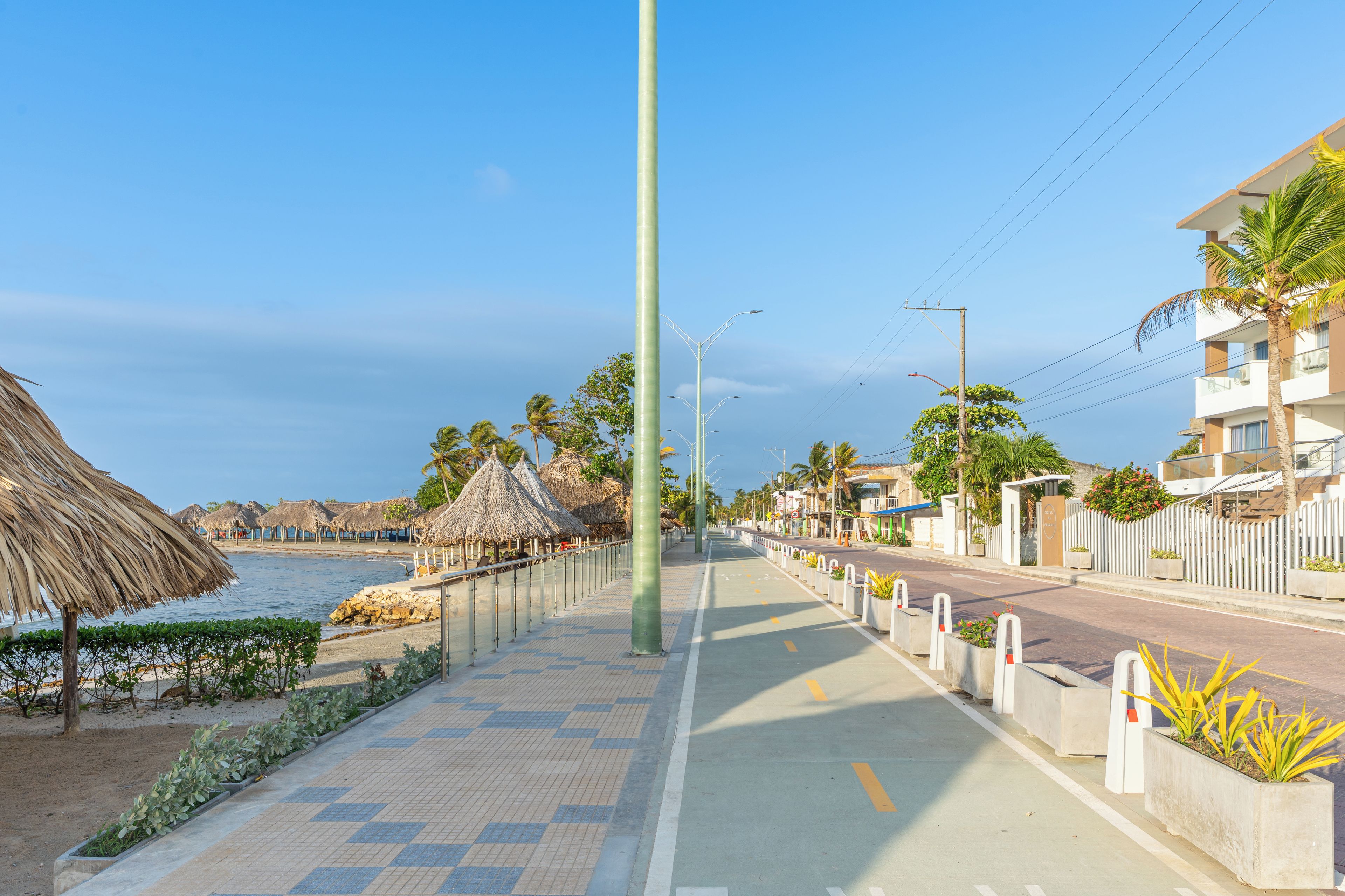 Una passeggiata costiera con una spiaggia da un lato, palme e una strada con edifici e una recinzione.
