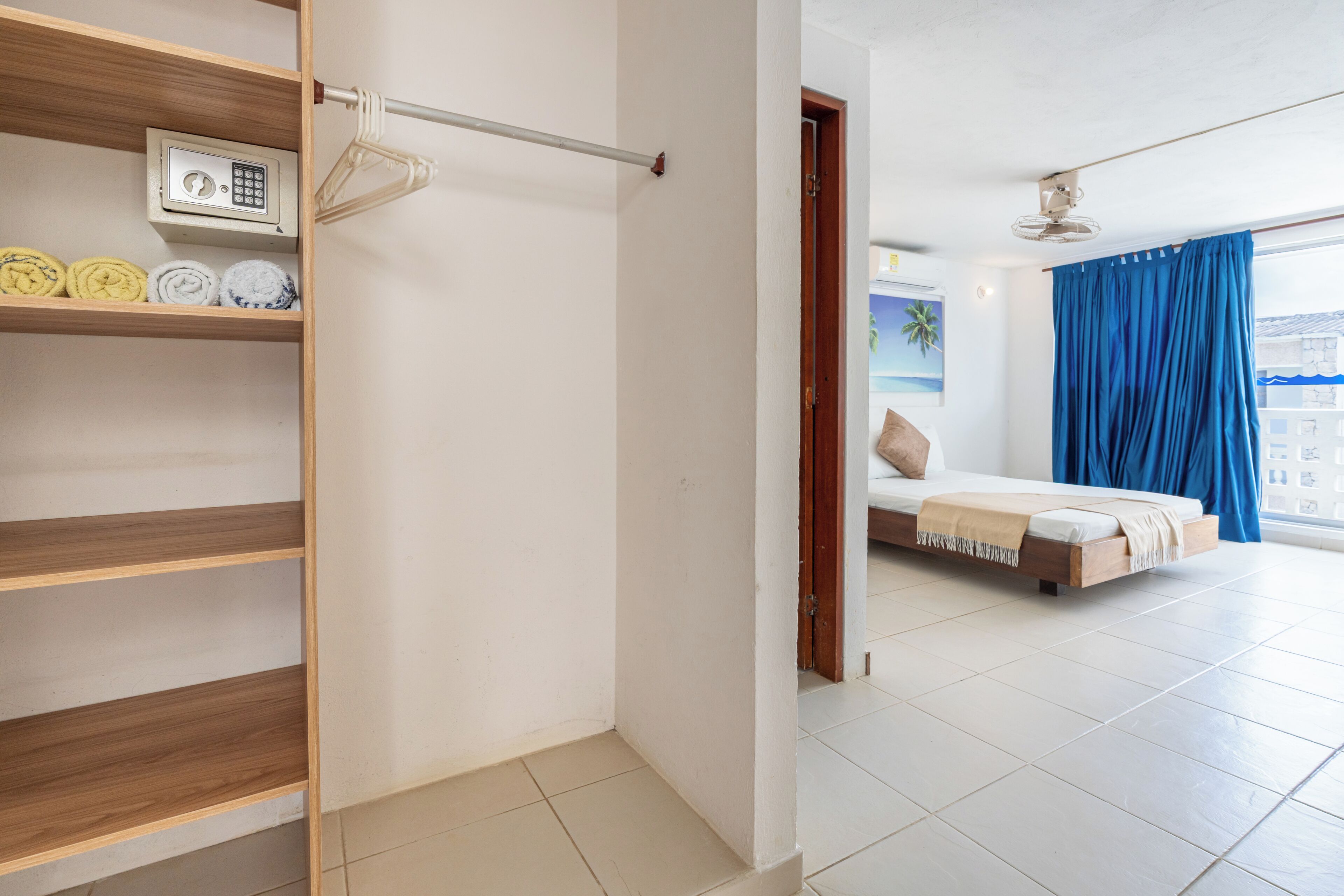 A bedroom with a bed, a wooden shelf with towels, a security device, and a view of the outside.