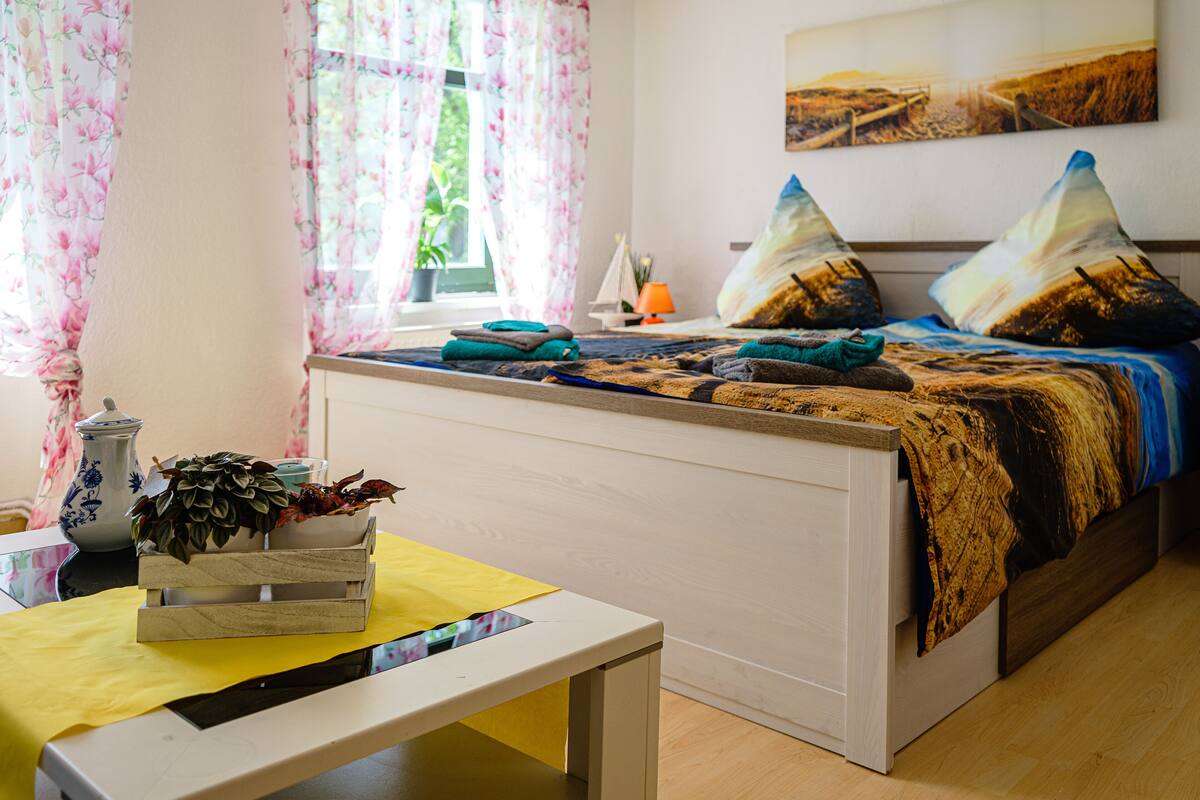 Quarto com cama, mesa de cabeceira, janela com cortinas, planta em vaso e um quadro na parede.