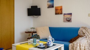 A living room with a blue sofa, a white coffee table with a yellow tablecloth, a TV mounted on the wall, and three framed pictures. - Nordstern Restaurant & Pension (Gingst)