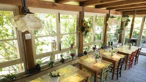 Un restaurant avec de nombreuses tables dressées pour le service, de grandes fenêtres ornées de plantes et un sol carrelé.