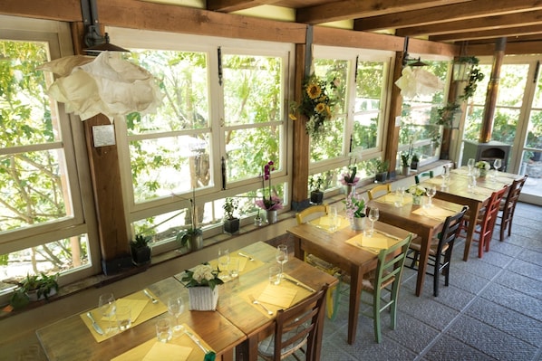 A restaurant with multiple tables set for dining, large windows with plants, and a tiled floor.