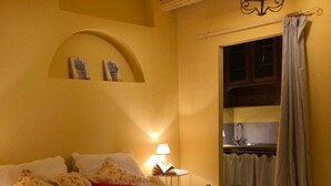 Une chambre confortable avec un lit, des oreillers, une table de chevet avec une lampe, une petite commode en bois et une vue sur une cuisine par une porte ouverte.