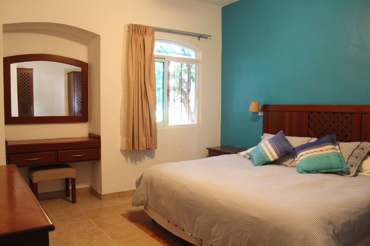 A bedroom with a bed, a desk, a chair, a mirror, and a window with curtains.