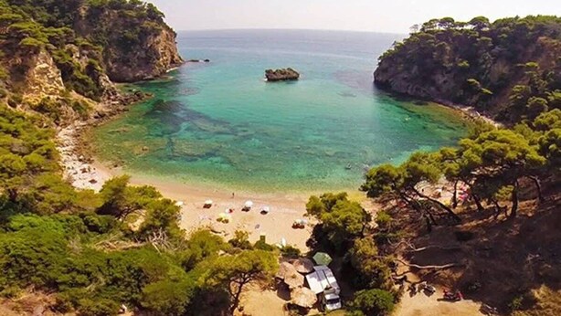 A coastal landscape with clear turquoise waters, rocky cliffs, and a beach area with sparse vegetation and a few structures.