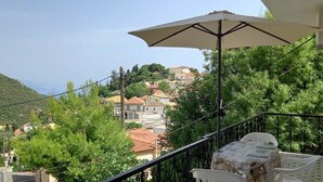 A balcony with a white chair and a table, overlooking a hillside with houses and greenery.