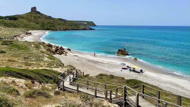 Paisaje costero con playa, una escalera de madera que baja a la playa y una colina con una estructura en la cima.