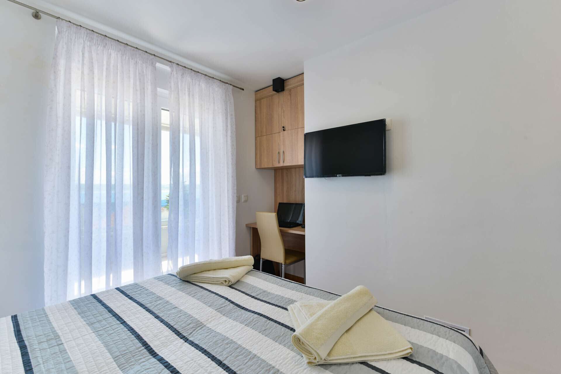 Ein Hotelzimmer mit Bett, Schreibtisch mit Stuhl, Wandfernseher und Fenster mit leichtem Vorhang.