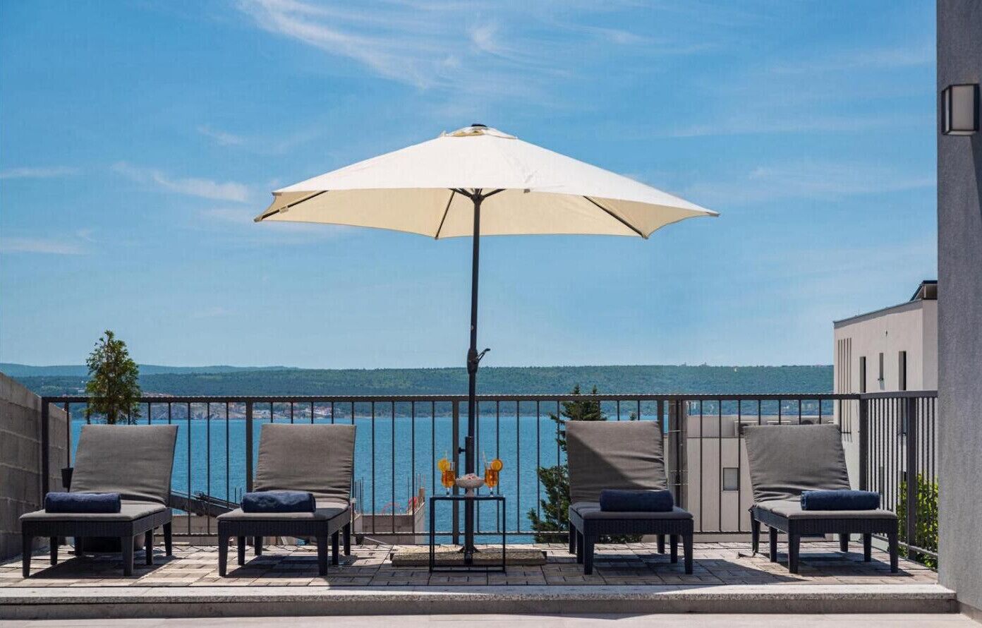 Ein Balkon mit vier Liegestühlen, einem Tisch mit einer Vase und zwei Flaschen sowie einem großen Sonnenschirm.