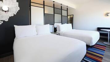 Quarto de hotel com duas camas, uma mesa pequena, uma cadeira e um tapete listrado e colorido.