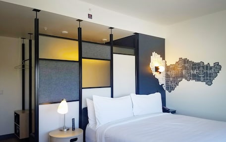 A modern hotel room with a bed, a bedside table with a lamp, a wall-mounted light fixture, and a hanging light fixture.