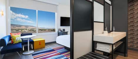 A modern hotel room with a large window, a bed, a desk, a bathroom, and a colorful striped rug.