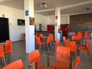 A spacious dining area with orange chairs and tables, a kitchen area with red cabinets, and a stone wall.