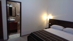 A hotel room with a bed, a nightstand, a wall-mounted light, a mirror, and a bathroom with a sink and mirror.