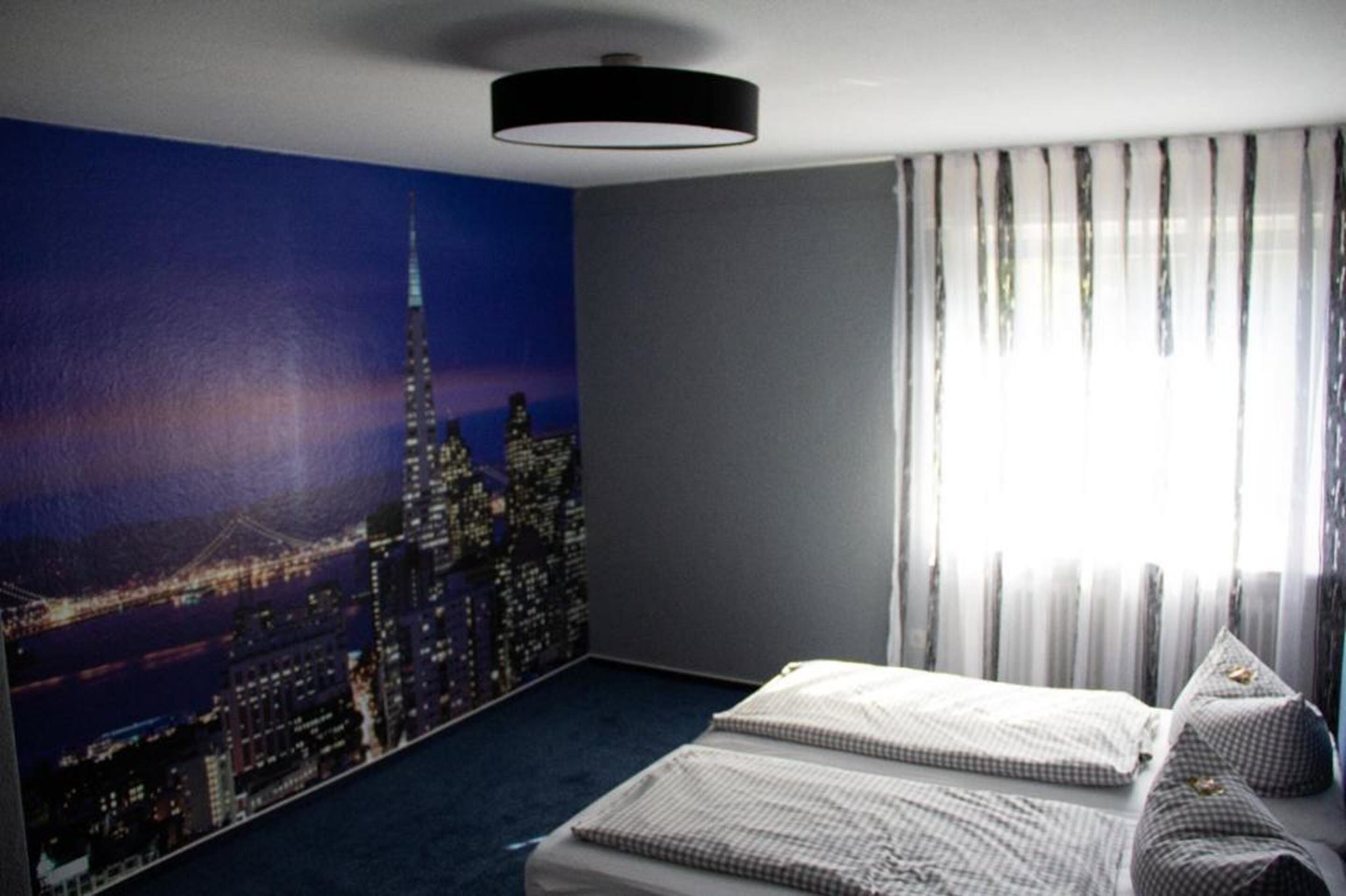 A bedroom with a cityscape mural, a bed with checkered bedding, and a large window with sheer curtains.
