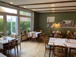 A dining area with tables set for four, wooden chairs, and a view through large windows. - Stadthotel im Kolpinghaus Lingen (Lingen)