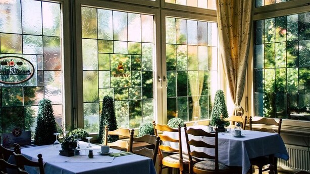 A dining area with tables set for two, large windows with frosted glass, and a view of greenery outside. - Hotel Kolpinghaus Lingen (Lingen)