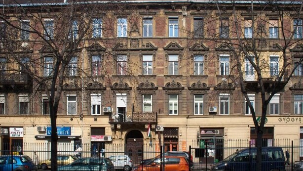 Un edificio de varios pisos con estilo arquitectónico clásico, que presenta una entrada central con un arco decorativo y una bandera. Hay varios coches aparcados en la parte delantera, y el edificio cuenta con varios espacios comerciales a nivel de calle con señalización visible.