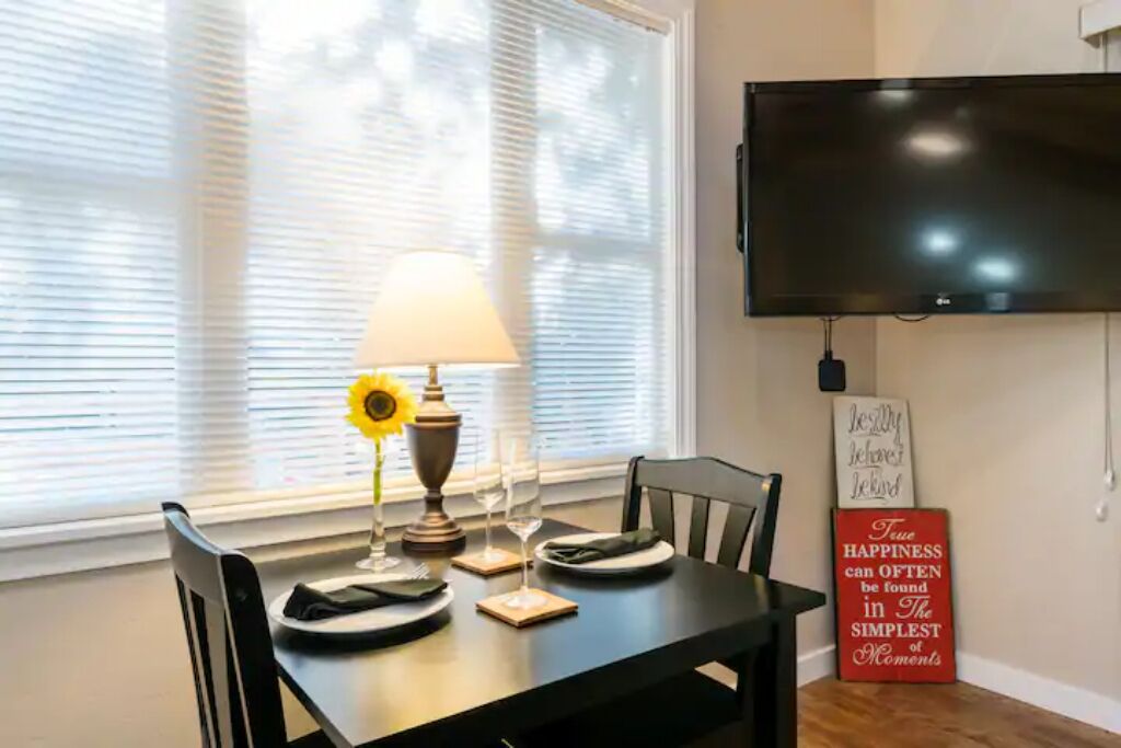A dining area with a table set for two, a lamp, and a sunflower centerpiece. A flat-screen TV is mounted on the wall above the table.
