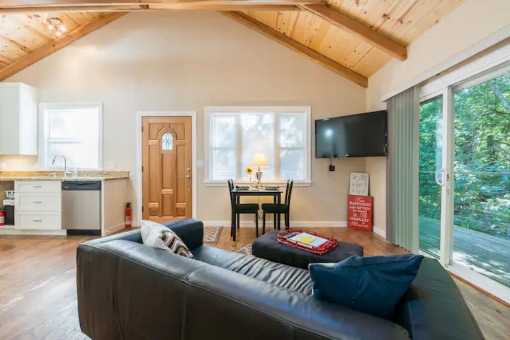 A cozy living room with a black leather sofa, a dining table with chairs, a wooden ceiling, and a large window with a view of greenery.