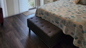 Un dormitorio con cama, un banco, un ventilador de techo, una mesita de noche, una lámpara, un cuadro y vistas al exterior.