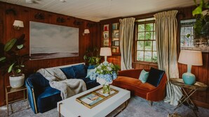 A cozy living room with a blue sofa, an orange armchair, and a white coffee table. There is a large painting on the wall, a potted plant, and a window with curtains. - Historic Arcadia: Heritage Coachman Lodge on 2000-acre Seaside Estate (Portpatrick)