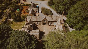 An aerial view of a historic stone building with multiple chimneys, surrounded by dense greenery and a winding road. - Historic Arcadia: Heritage Coachman Lodge on 2000-acre Seaside Estate (Portpatrick)