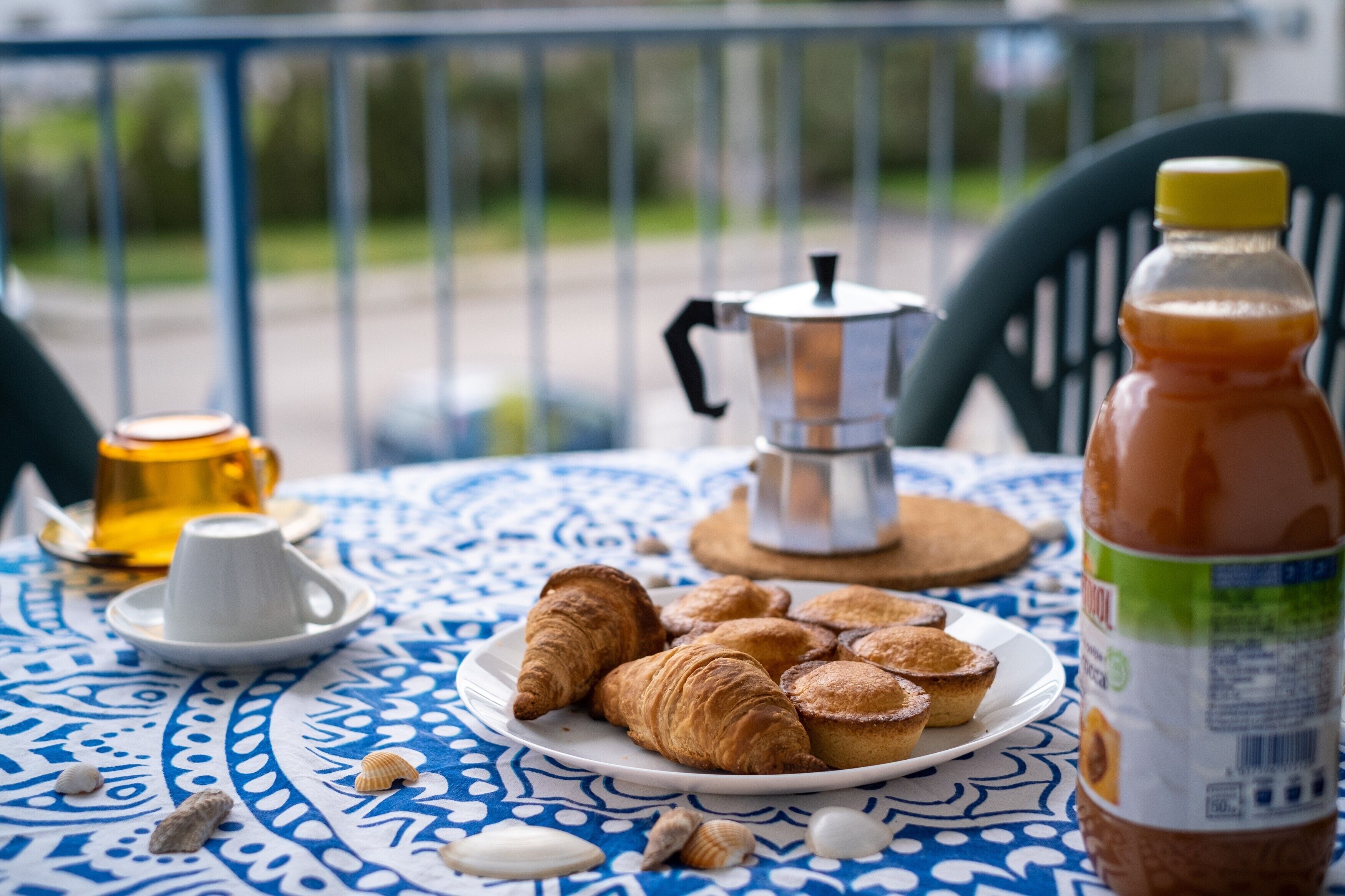 Un tavolo con un piatto di croissant, una caffettiera, una bottiglia di succo e una tazza di caffè su una tovaglia fantasia.