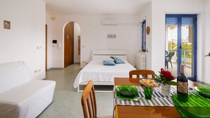A bedroom with a bed, a dining table with chairs, a balcony with a chair, and a door leading to another room.